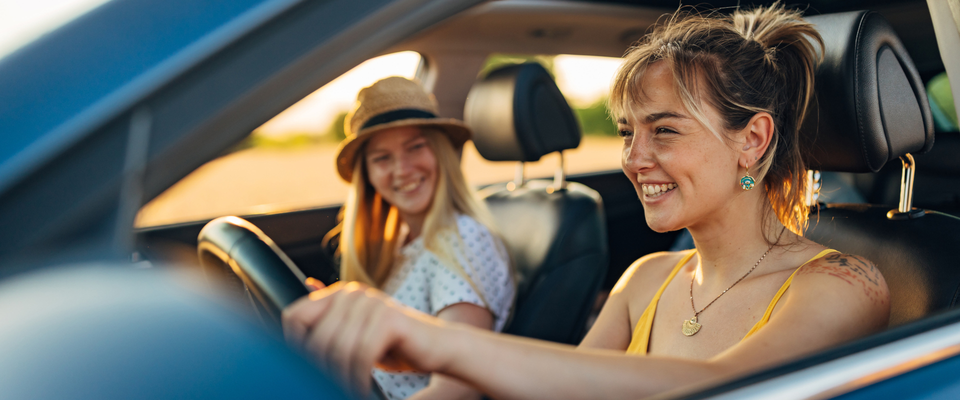 Two women smiling in a car, looking directly at the camera with cheerful expressions.